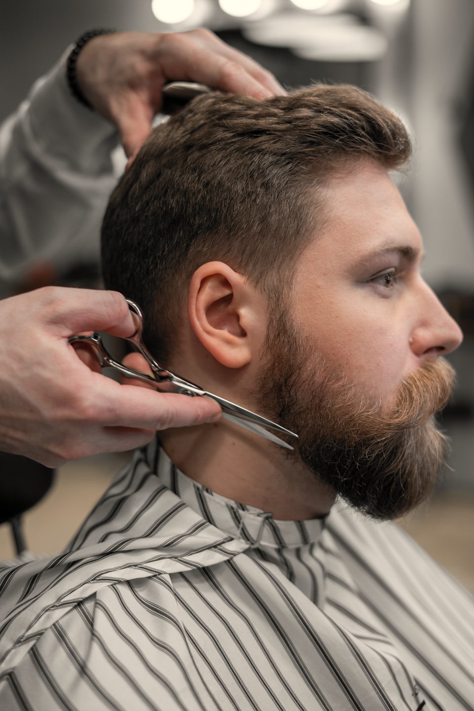 Barbiere che taglia la barba rossa di un uomo con mantellina a righe.